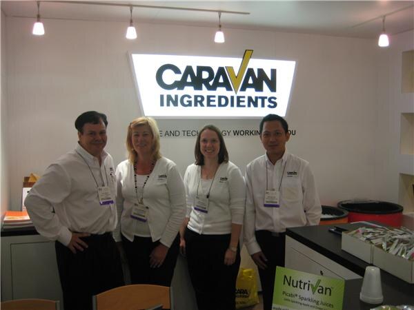 Two men and two women in white shirts and lanyards stand in front of an illuminated Caravan Ingredients sign, with a Nutrivan Picabi sparkling juices sign on the counter.
