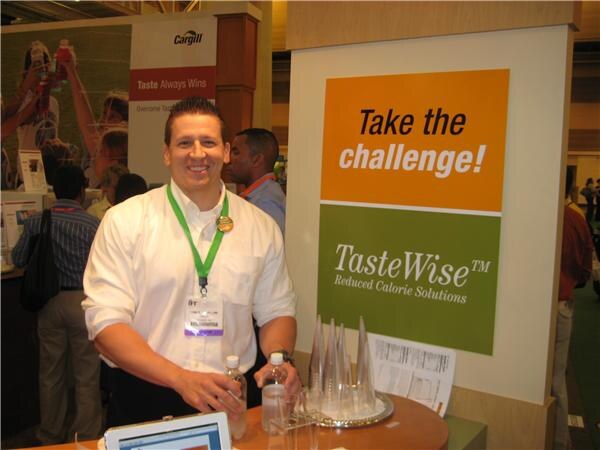 A smiling man in a white shirt and green lanyard stands at a Cargill booth beside a TasteWise sign reading Take the challenge, with water bottles and glasses on the table.