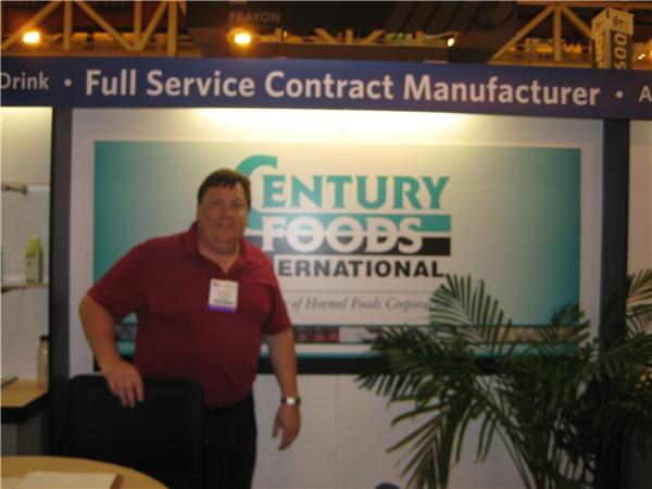 A man in a maroon shirt stands in front of a Century Foods International sign reading Full Service Contract Manufacturer, next to a potted palm.
