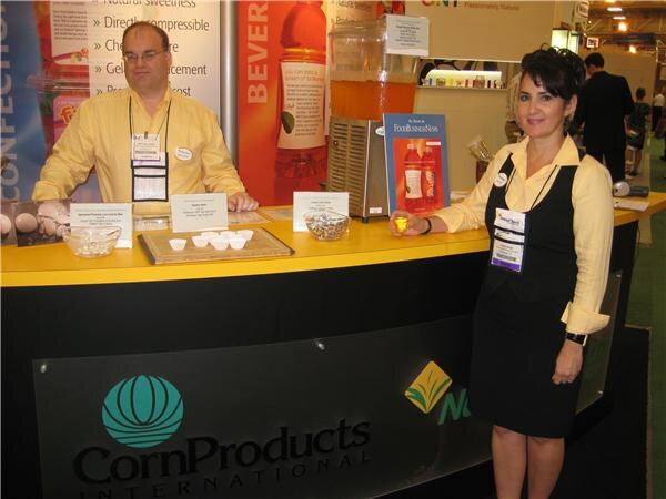 A man and a woman in yellow shirts stand behind a yellow and black counter at the CornProducts International booth, with sample cups and a FoodBusinessNews display.