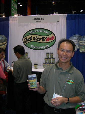 A smiling man in a green polo holds a small jar at the JavaVoo booth, with a JavaVoo Micro-Brew Technology sign and more jars on a table behind him.