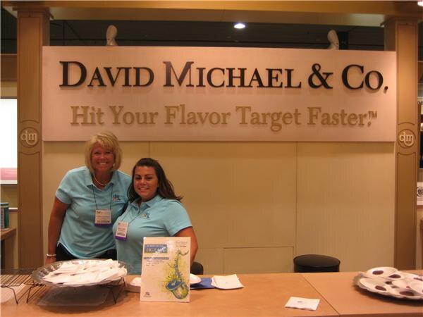 Two women in light blue polo shirts stand behind a counter under a David Michael & Co. sign reading Hit Your Flavor Target Faster, with sample plates in front of them.