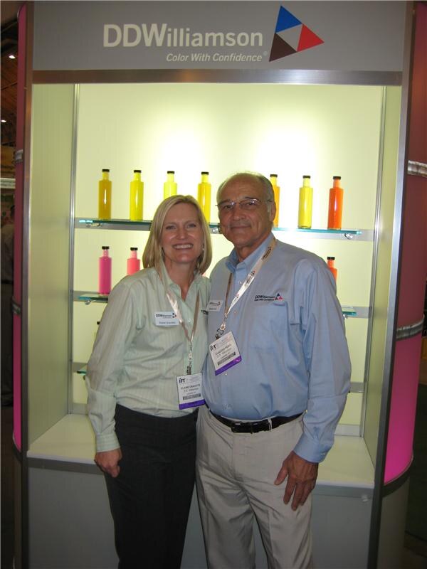 A woman and a man wearing IFT badges stand in front of a DDWilliamson display case holding bottles of yellow, orange and pink liquid.