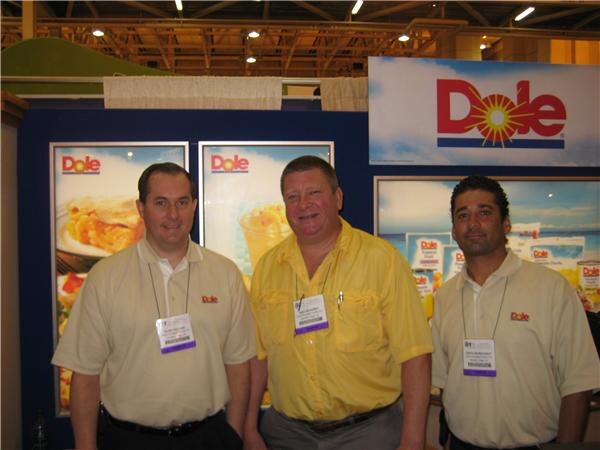 Three smiling men in tan and yellow shirts with show badges stand in front of the Dole booth, which has Dole logo signs and photos of food and fruit cups.