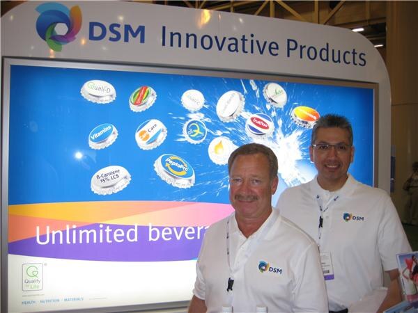 Two men in white DSM polo shirts stand in front of a lit DSM Innovative Products display showing bottle caps and the partial text Unlimited bever.