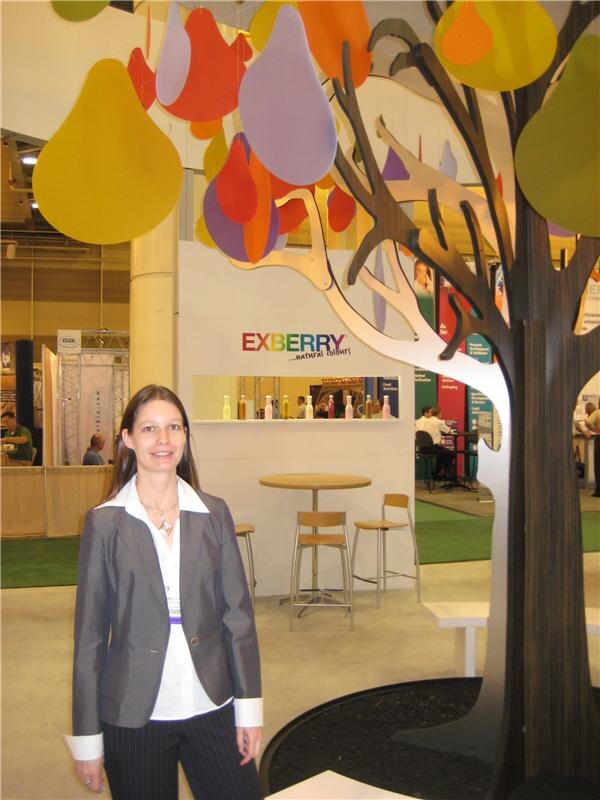 A smiling woman in a gray blazer stands in front of the Exberry natural colours booth, which has a decorative tree and colorful hanging teardrop shapes.