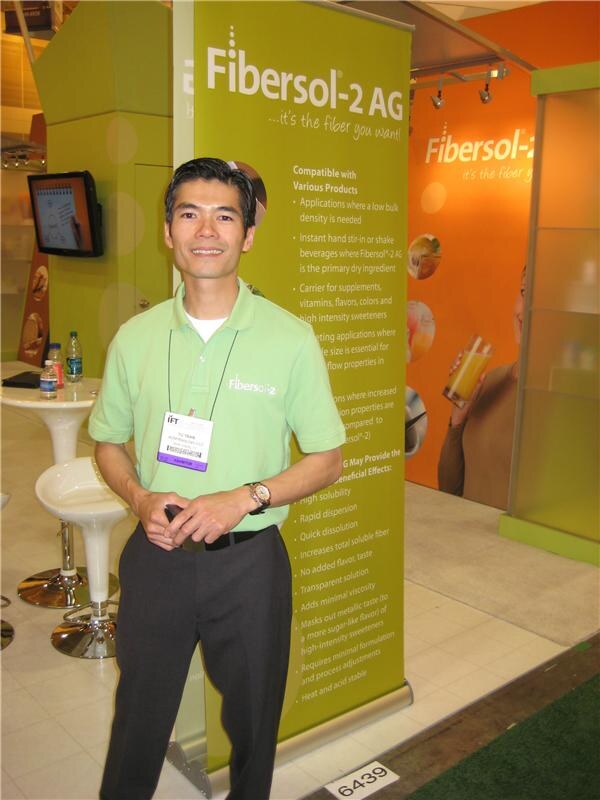 A smiling man in a light green Fibersol polo and show badge stands beside a green Fibersol-2 AG banner in an orange and green booth with a white stool and a wall-mounted screen.