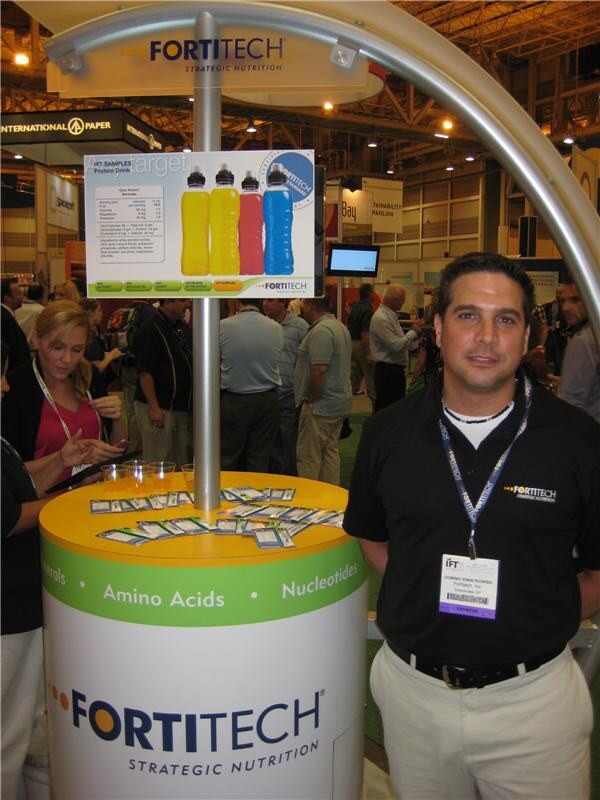 A man in a black Fortitech polo and exhibitor badge stands beside a Fortitech Strategic Nutrition counter with sample cups, handouts and a sign showing four colored beverage bottles.