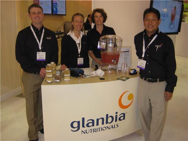 Four people in black shirts with badges stand behind a Glanbia Nutritionals counter holding a dispenser of red drink and small sample jars.
