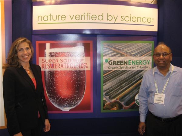 A smiling woman in a dark blazer and a man in a blue shirt with a badge stand beside displays for Super Soluble Resveratrol 10% and GreenEnergy Organic Spirulina and Chlorella.
