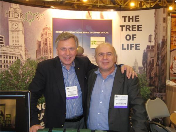 Two men in dark jackets stand with arms around each other's shoulders in front of a Hidrox banner reading The Tree of Life, both wearing IFT badges.