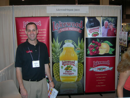 A smiling man in a black polo with a name badge stands in front of Lakewood Organic Juices banners showing a bottle of Pure Pineapple juice and cranberry juice graphics.