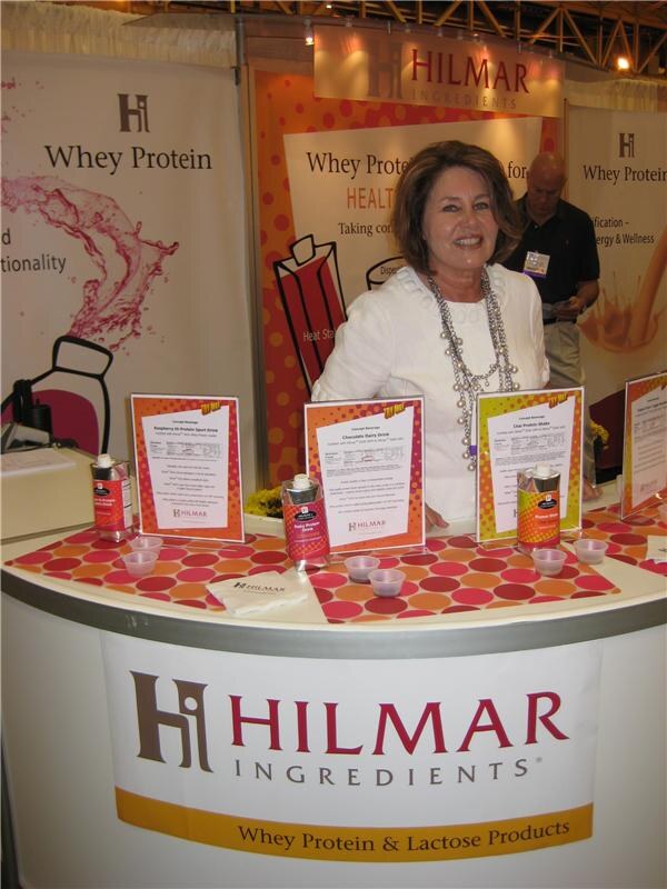 A smiling woman in white stands behind a curved Hilmar Ingredients counter displaying whey protein beverage sample cards, small cartons and sample cups.