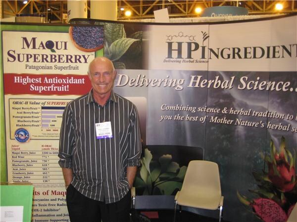 A smiling bald man in a striped shirt with a badge stands in front of HP Ingredients and Maqui Superberry banners, including an antioxidant chart.