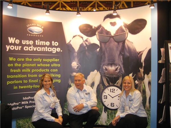 Two women and a man in light blue shirts pose in front of an Idaho Milk Products banner showing a cow wearing sunglasses and a clock, with the text We use time to your advantage.