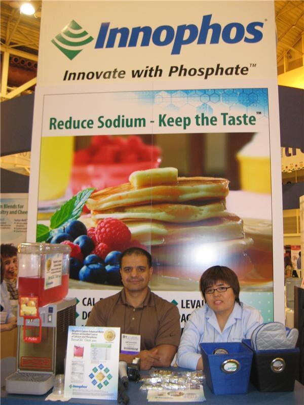 Two seated people, a man in a brown shirt and a woman with glasses in a light blue shirt, at the Innophos booth under a sign reading Reduce Sodium - Keep the Taste, beside a beverage dispenser.