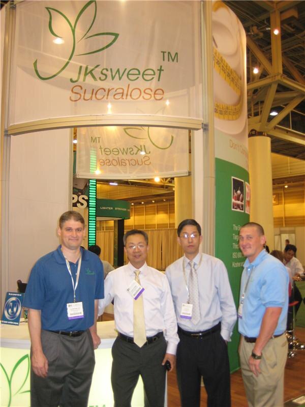 Four men with show badges stand together in front of the JKsweet Sucralose booth, two in blue shirts and two in white dress shirts with ties.