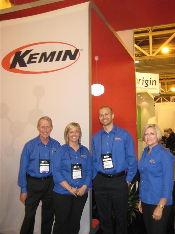 Two men and two women in matching blue shirts and badges stand in front of the red and white Kemin booth, with an Origin sign visible in the background.