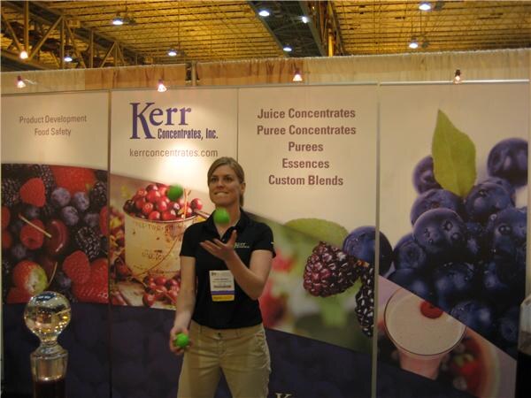 A woman in a black shirt juggles small green balls in front of Kerr Concentrates, Inc. banners listing juice concentrates, purees, essences and custom blends, with berry images.