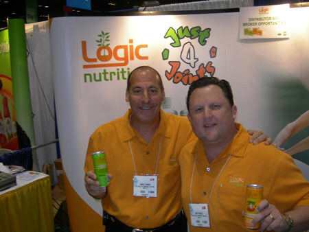 Two men in orange shirts hold cans in front of a Logic Nutrition and Just 4 Joints display.