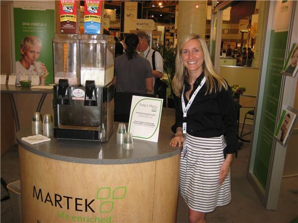 A smiling woman with long blonde hair stands beside a Martek booth counter holding a beverage dispenser, with Horizon Organic cartons above it and a Today's Menu sign.