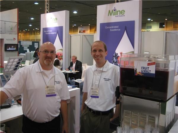 Two smiling men in white polo shirts and show badges stand in the Milne Fruit Products booth beside a beverage dispenser labeled Fruit & Malt, with Milne banners behind them.