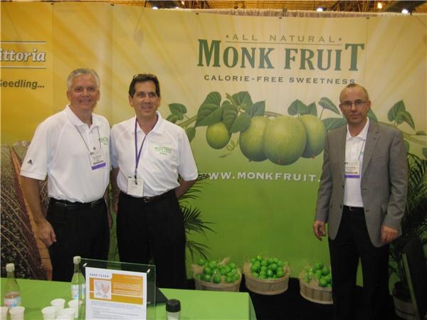 Three men in a booth stand in front of a green Monk Fruit backdrop reading "All Natural Monk Fruit, Calorie-Free Sweetness." Baskets of green fruit sit on the table in front of them.