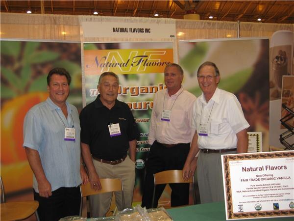Four men with show badges stand together at the Natural Flavors Inc booth, with a sign about Fair Trade organic vanilla in the lower right corner.