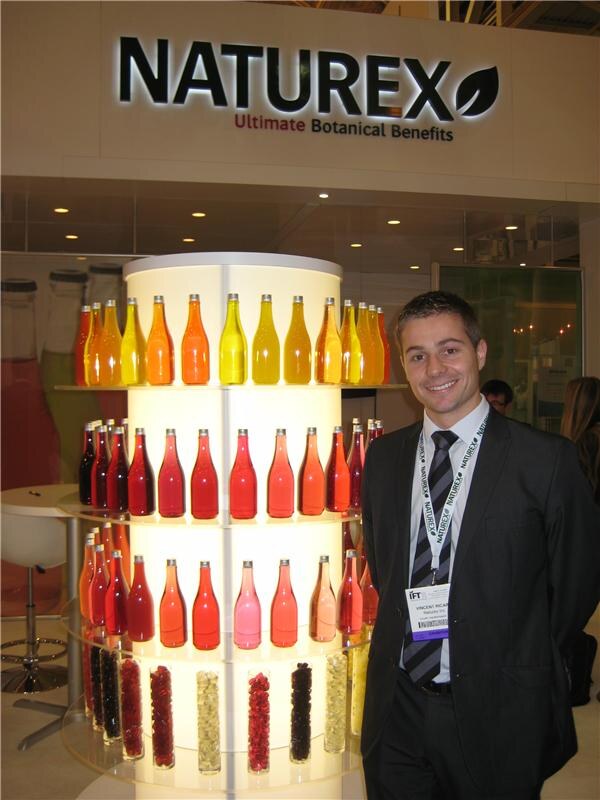 A smiling man in a dark suit and striped tie with a Naturex lanyard stands beside a lit display of colorful bottles under a Naturex sign reading Ultimate Botanical Benefits.