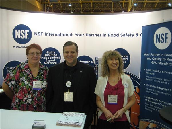 Two women and a man stand behind a counter at the NSF International booth, with banners about food safety and quality behind them.