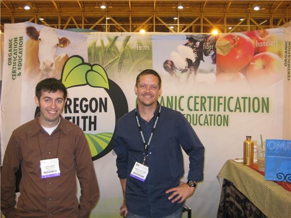 Two smiling men with badges stand in front of an Oregon Tilth banner about organic certification and education, showing a cow, apples and crops.