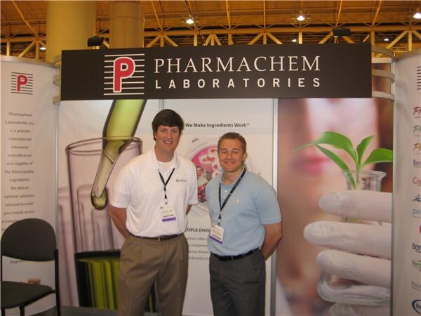 Two men in polo shirts with badges stand in front of the Pharmachem Laboratories booth sign and graphics showing a dropper and a seedling.
