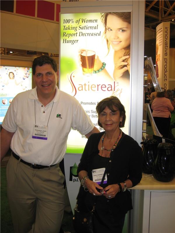 A man in a white polo shirt and a woman in a black cardigan stand together in front of a lit Satiereal sign reading 100% of Women Taking Satiereal Report Decreased Hunger, at the P.L. Thomas booth.