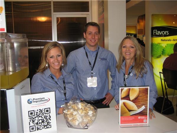 Two women and a man in blue striped shirts stand behind a counter at the Premium Ingredients booth, with a bowl of fortune cookies and a sign with a QR code in front of them.