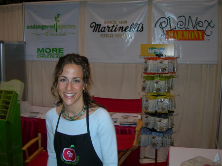 A smiling woman with dark curly hair and a black apron stands in a booth. Banners behind her read Endangered Species Chocolate, Martinelli's Gold Medal and Planex Harmony, with a rack of Harmony products.