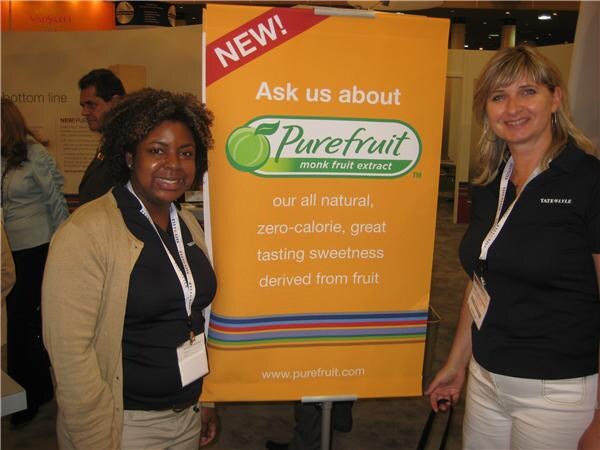 Two smiling women in dark Tate & Lyle polo shirts and lanyards stand beside an orange banner reading "NEW! Ask us about Purefruit monk fruit extract" at a trade show booth.