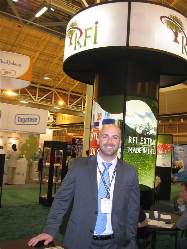 A smiling bald man in a gray suit and badge lanyard stands beside a round RFI display reading RFI Extract made in the USA. Tagatose and Lactalis signs appear in the background.