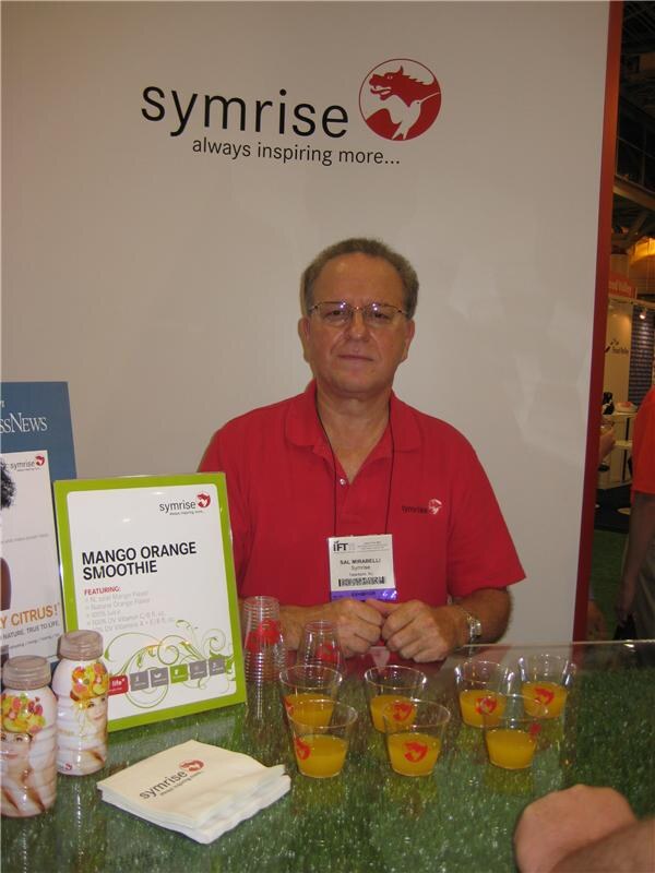Sal Mirabelli of Symrise, wearing glasses and a red polo, sits at a counter at the Symrise booth with cups of orange drink, a Mango Orange Smoothie sign and bottled smoothies.