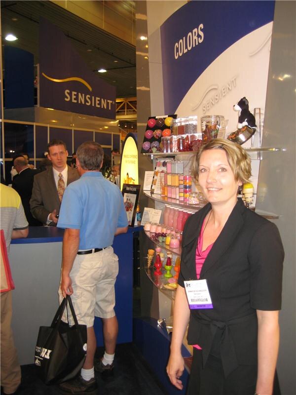 A smiling woman in a black jacket stands beside a Sensient Colors display shelf holding colorful candies and confections. Other attendees talk at the Sensient booth counter behind her.
