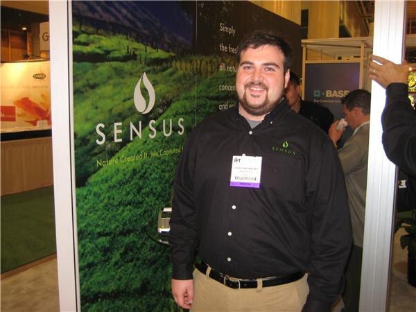 A smiling bearded man in a black shirt with a Sensus logo and a badge stands in front of a green landscape Sensus wall graphic. A BASF sign is visible in the background.