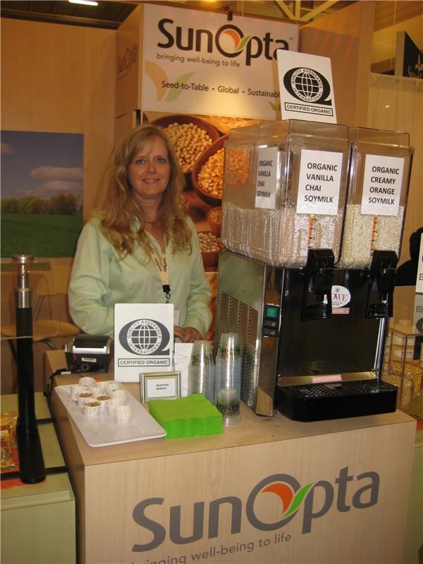 A woman with long blonde hair stands behind a SunOpta counter with two soymilk dispensers labeled Organic Vanilla Chai and Organic Creamy Orange, with Quality Assurance International certified organic signs.