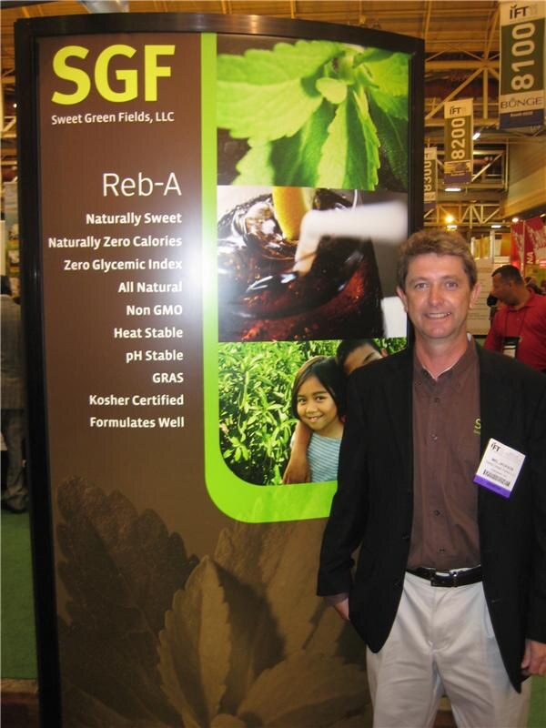 A smiling man in a dark jacket stands beside a Sweet Green Fields (SGF) Reb-A display listing product attributes, with stevia leaf images. A Bunge banner hangs overhead in the background.