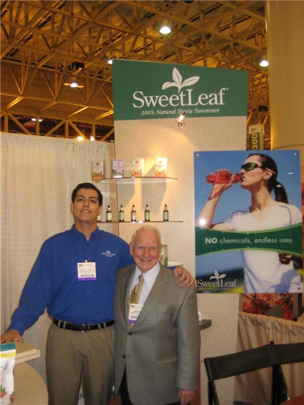 A younger man in a blue shirt stands with his arm around an older man in a gray blazer in front of a SweetLeaf stevia sweetener booth with product shelves and a poster of a woman drinking.