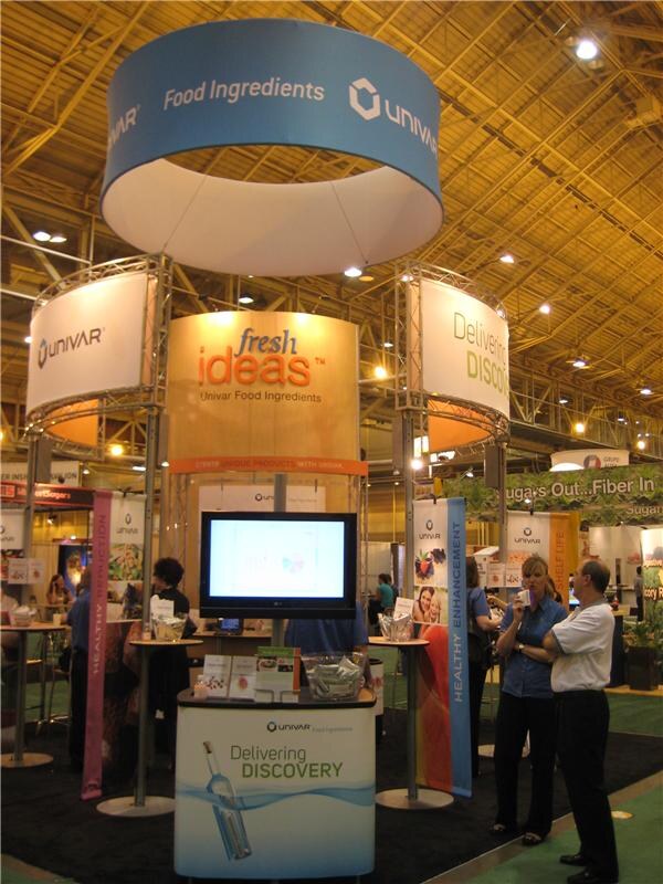 A large Univar Food Ingredients exhibit with a round overhead sign, a "fresh ideas" display and a video screen. Two people stand talking at the right side of the booth.