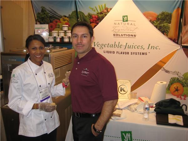 A smiling woman in a chef's coat and a man in a maroon polo stand at a Vegetable Juices, Inc. booth. Signs read Natural Ingredient Solutions and Liquid Flavor Systems.