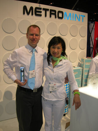 A man in a white shirt and blue patterned tie holds a small bottle beside a smiling woman in white, in front of a Metromint sign and stacked boxes.