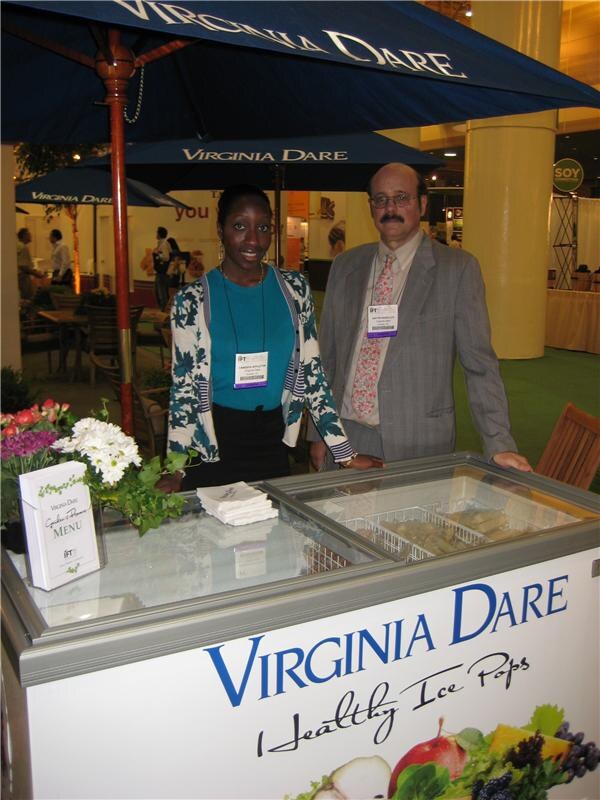 A woman and a man stand behind a freezer case marked "Virginia Dare Healthy Ice Pops" under blue Virginia Dare umbrellas. Flowers and a menu card sit on the left.