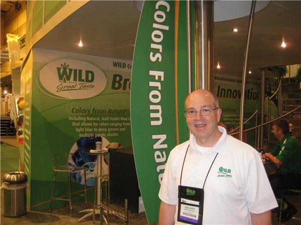 A smiling man with glasses and a white Wild polo shirt stands in front of a green Wild booth. Signs mention colors from nature and innovation, and other people sit in the background.