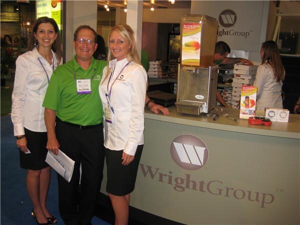 A man in a green polo and two women in white blouses stand beside the Wright Group booth counter. A beverage dispenser and product literature sit on the counter.