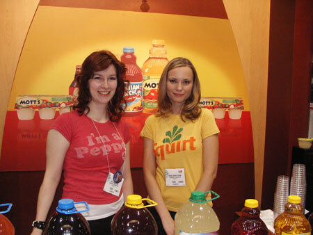 Two young women stand behind a counter lined with jugs of juice and soda, one in a red T-shirt and one in a yellow Squirt T-shirt, in front of a Mott's graphic.